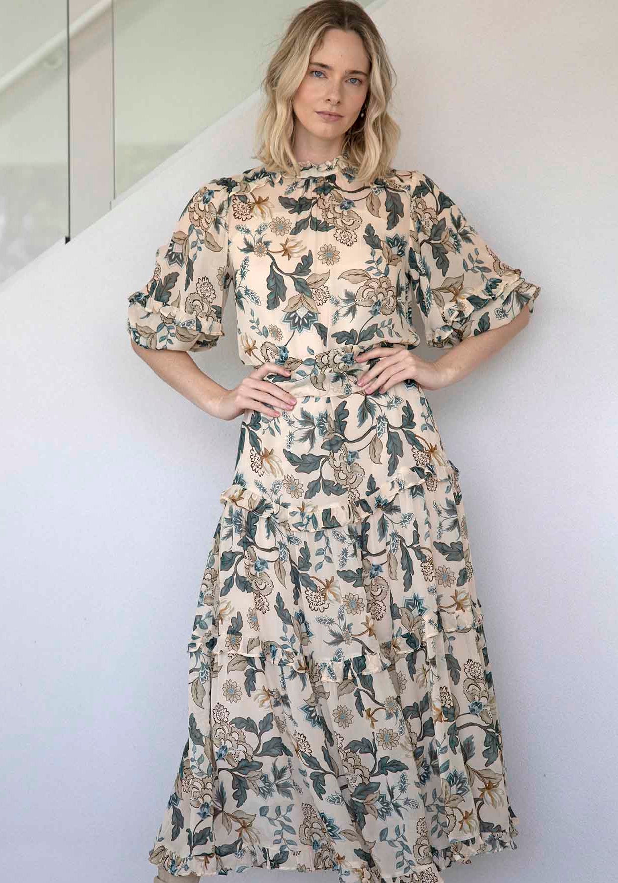 A woman modeling a beige midi dress with a dark green and brown botanical leaf pattern, featuring puff sleeves and a tiered skirt, paired with beige ankle boots against a white wall.